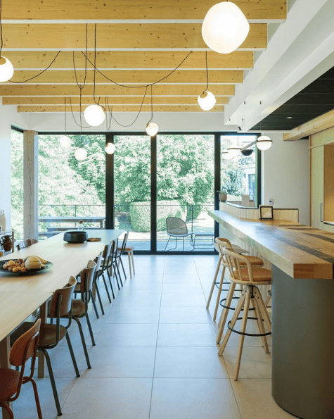 Salle lumineuse avec table de repas, cuisine moderne et grandes baies vitrées offrant une vue sur la nature.